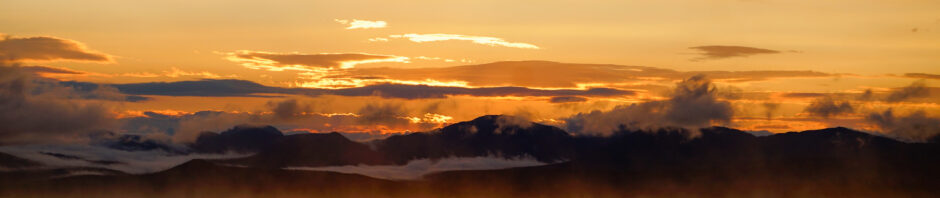 Golden sunset over calm water with distant mountains and low misty clouds in the distance.