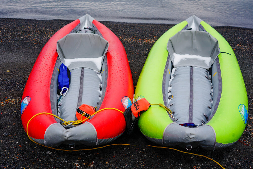 Two colorful inflatable kayaks (red and lime green) with grey seating sit on a pebbled beach by the water, with gear and ropes nearby.